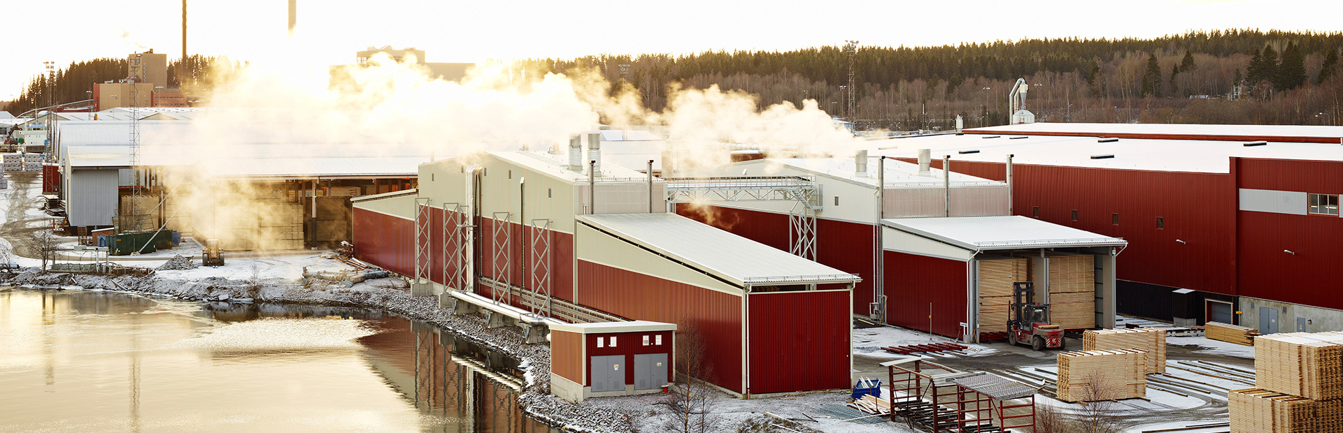 Röda Valutec kanaltorkar TC i vintermiljö vid vatten. Ånga stiger från taken och virke hanteras med truck framför torkarna.