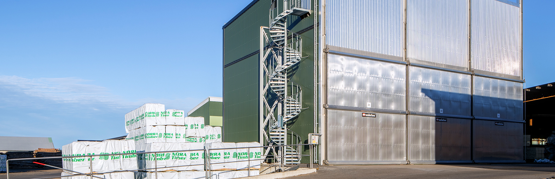 Valutec truckmatad kammartork med spiraltrappa på fasaden och staplade Norra Timber-paket i förgrunden.